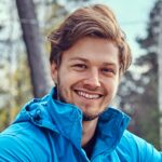 Portait photo d’un homme souriant face à la caméra, portant un manteau bleu, cheveux court au vent, fond en extérieur