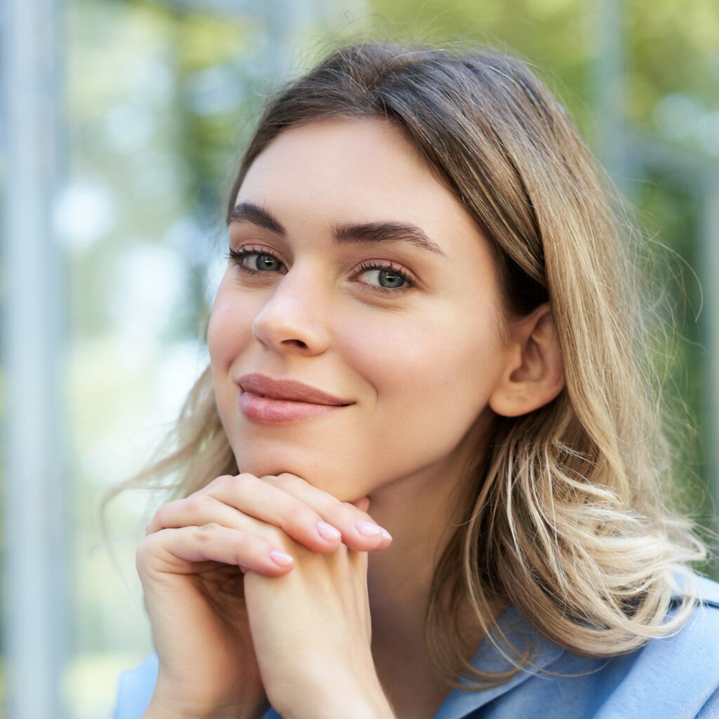 Portait photo d’une femme souriante, elle pose sa tête sur ses deux mains croisé, fond en extérieur flouté.