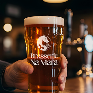 Mock up de bière avec le logo Na Mara imprimé en blanc sur le verre transparant. Une main attrape le verre. Fond de la photo en brasserie