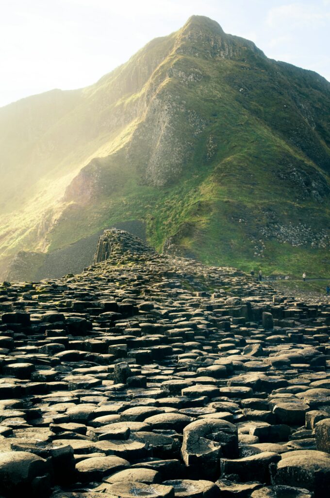 Photographie d’un paysage, roches cylindrique en premier plan, et une montagne verdoyante en fond. Lumière forte avec le soleil