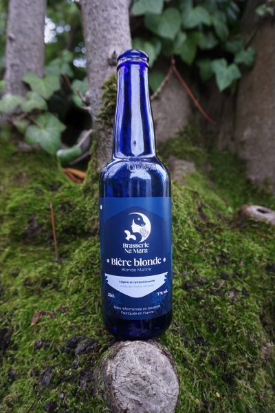 Photographie de produit : bouteille de bière Na Mara, parfum Blonde Marine. La bouteille bleue est posé sur un arbre moussus.